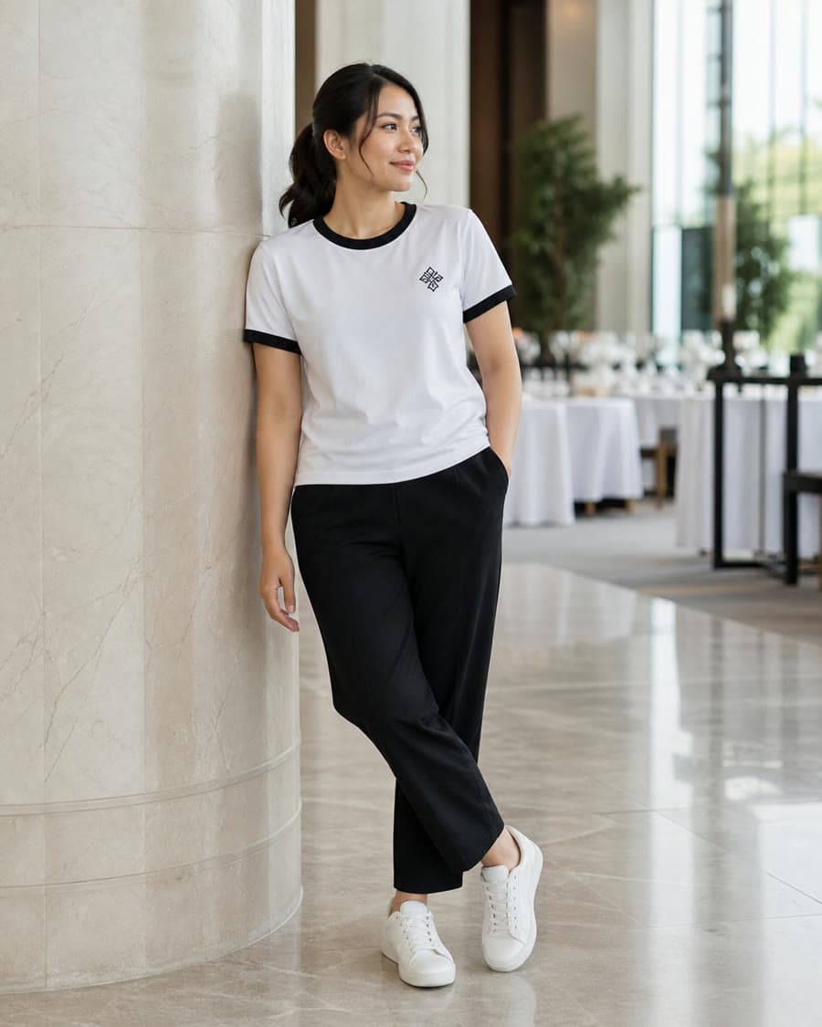 ผู้หญิงใส่เสื้อยืดสีขาว ขลิบคอและปลายแขนสีดำ ปักเล็กที่อกซ้าย ยืนพิงเสาในล็อบบี้